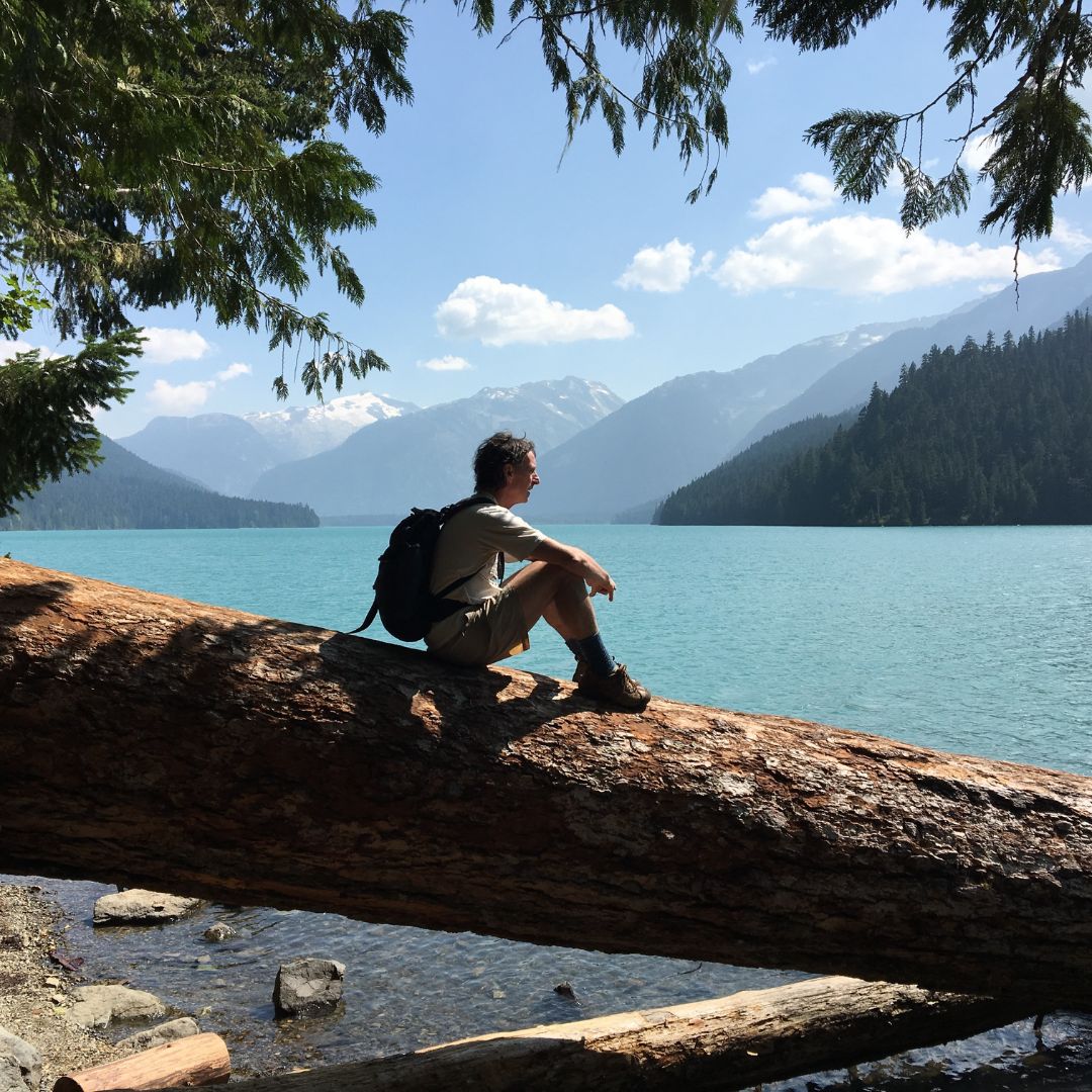 man sitting on fallen tree trunk looking out at a lake